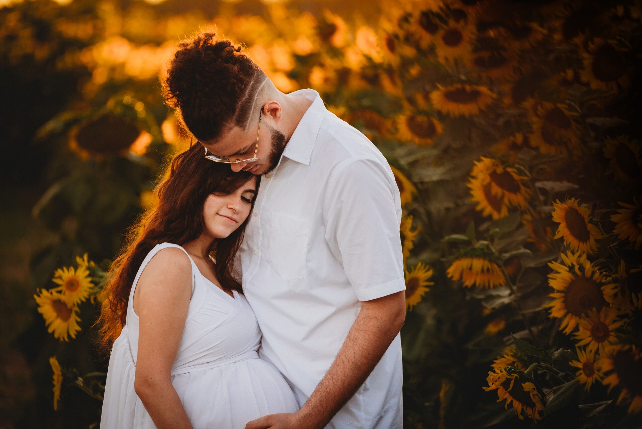 maternity couple in white in the sunflowers at sunset staring down