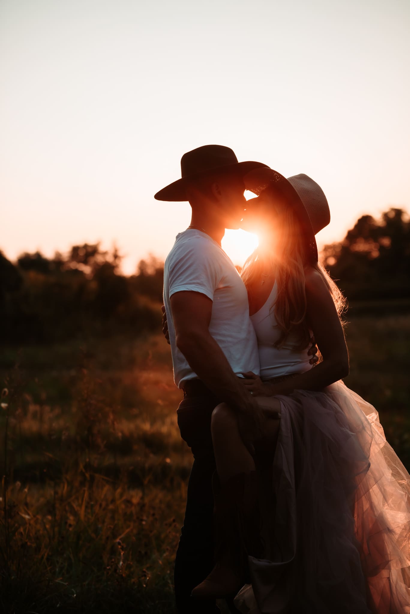 silhouette of man and woman kissing with the sun flaring between them with cowboy hats on