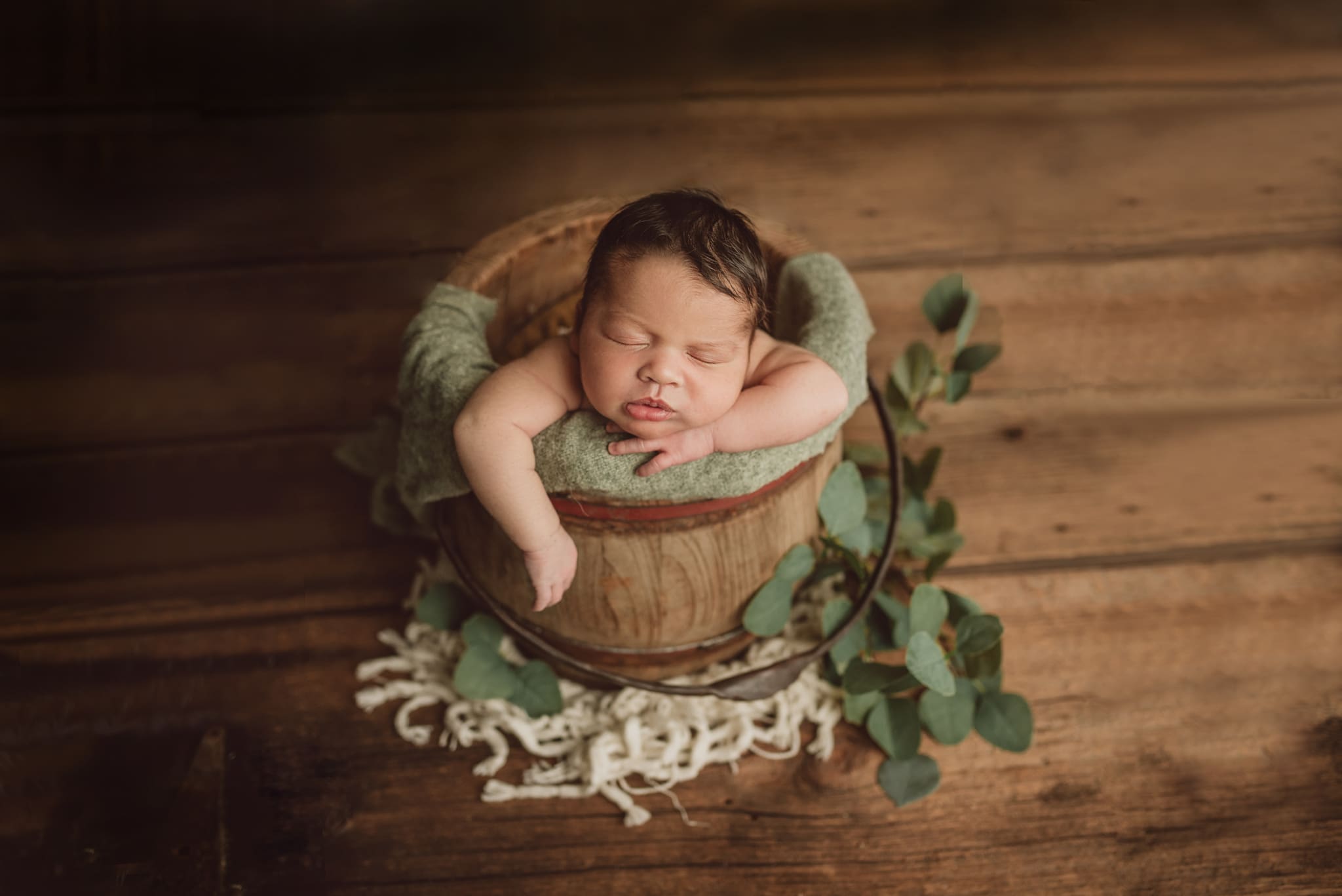 baby in bucket in sage color wrap with greenery around the bottom of the bucket