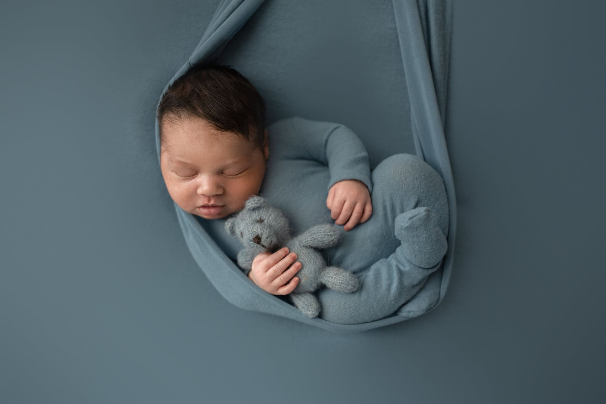 columbus ohio newborn photographer0050 baby boy laying on light blue fabric in a hammock style sling holding a knit teddy bear