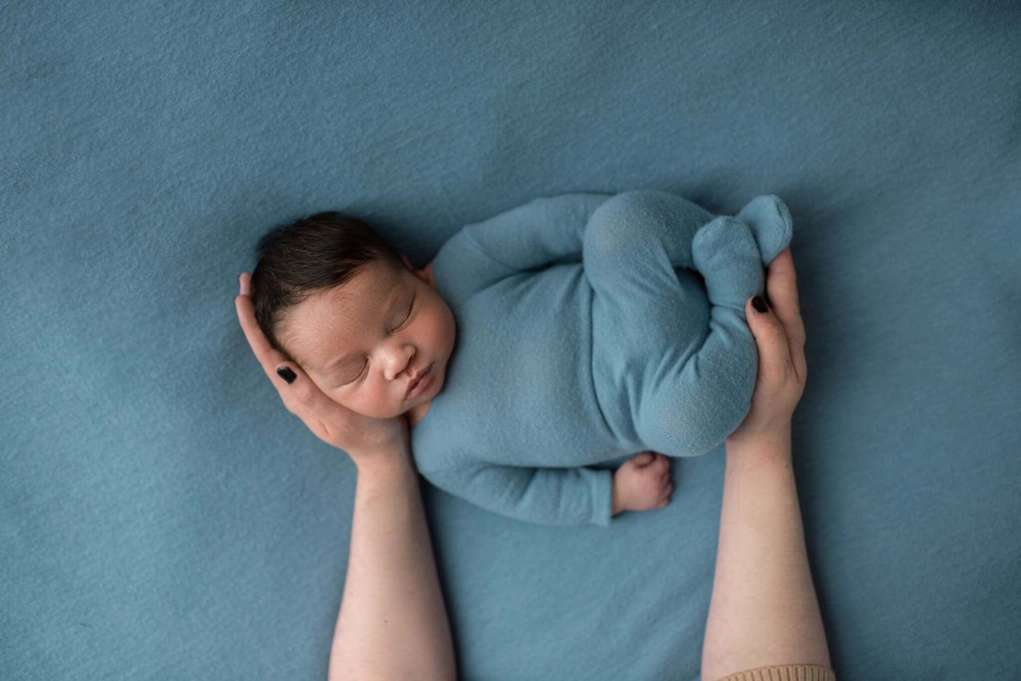 newborn boy on blue backdrop wearing a knit blue pj to match with mom's hands holding his head and feet